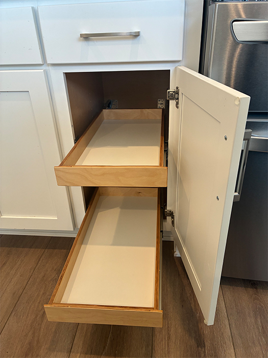 2-Single-Short-shelf-cabinet-A An open white kitchen cabinet reveals two wooden pull-out shelves, one positioned above the other, with a stainless steel appliance visible on the right and a wood floor below.