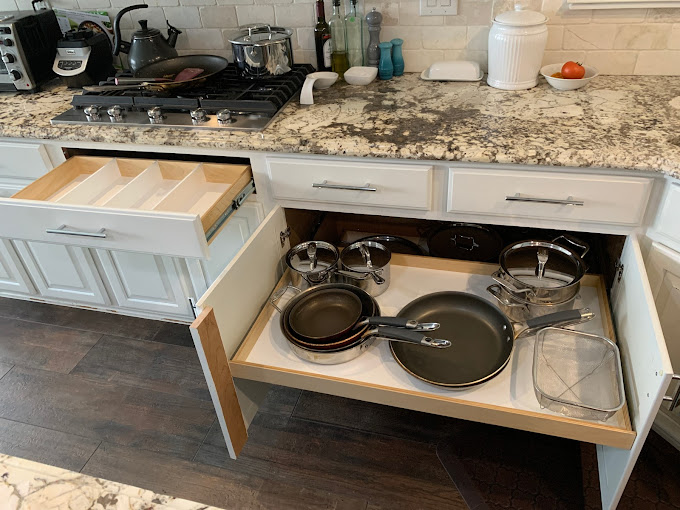 Glenn2020-08-20 An open kitchen drawer reveals pots, pans, and cookware neatly organized. Above it, another drawer with utensil trays is partially open. The countertop holds various kitchen items and appliances.