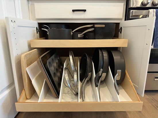 A kitchen drawer is open, revealing neatly organized cookware. The bottom shelf has baking sheets, pans, and lids separated by vertical dividers, while the top shelf holds pots and saucepans with handles facing outward.