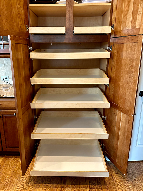 A wooden pantry cabinet with its doors open showing six pull-out shelves, all empty. The shelves are light-colored wood and slide out for easy access. The cabinet sits on a hardwood floor in a kitchen setting.