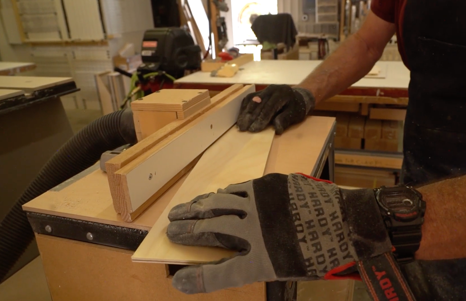 REOS workshop3 A person wearing work gloves guides a piece of wood through a table saw in a workshop, using a fence to ensure a straight cut. Tools and materials are visible in the background.