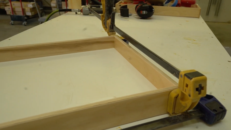 REOS workshop5 A wooden frame is being clamped together on a workbench, with glue visible at the joints. Tools and workshop items are seen in the background.