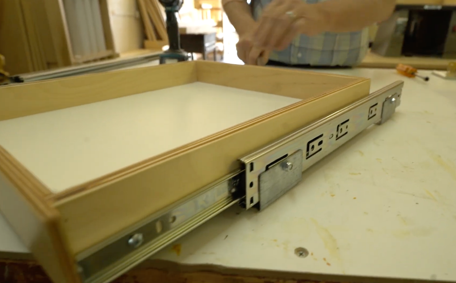 REOS workshop7 A person installs a drawer with metal slides and built-in electrical outlets on the side, in a woodworking shop. The drawer sits on a workbench among tools and wood pieces.