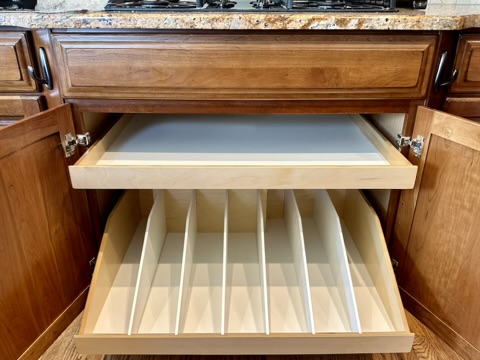 A kitchen cabinet with open doors shows two pull-out shelves; the top shelf is flat and empty, while the lower shelf has vertical dividers for organizing baking sheets or trays.