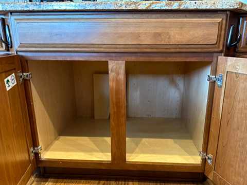 An empty wooden cabinet with two open doors is shown beneath a speckled countertop. The cabinet has a central divider and no shelves or items inside. The floor beneath is wood.