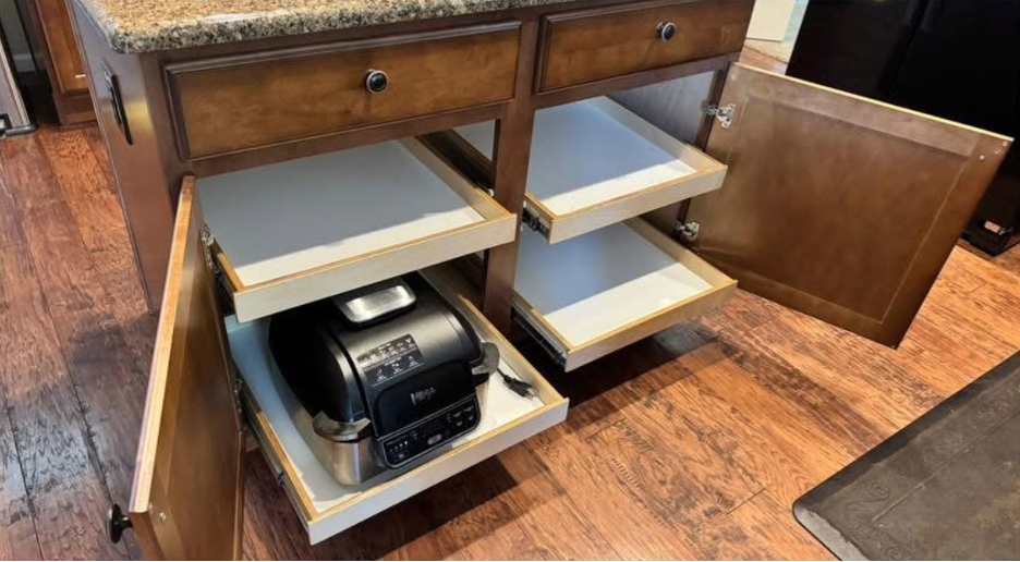 appliance reorg in island Brown kitchen cabinets with open doors reveal three pull-out shelves on each side. The bottom left shelf holds a black countertop appliance, while the others are empty. The cabinet is on a wood floor.