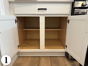 ba-1 A white kitchen cabinet with both doors open, revealing two empty interior sections separated by a vertical divider and a narrow wooden shelf inside. The number 1 is in a white circle at the bottom left corner.