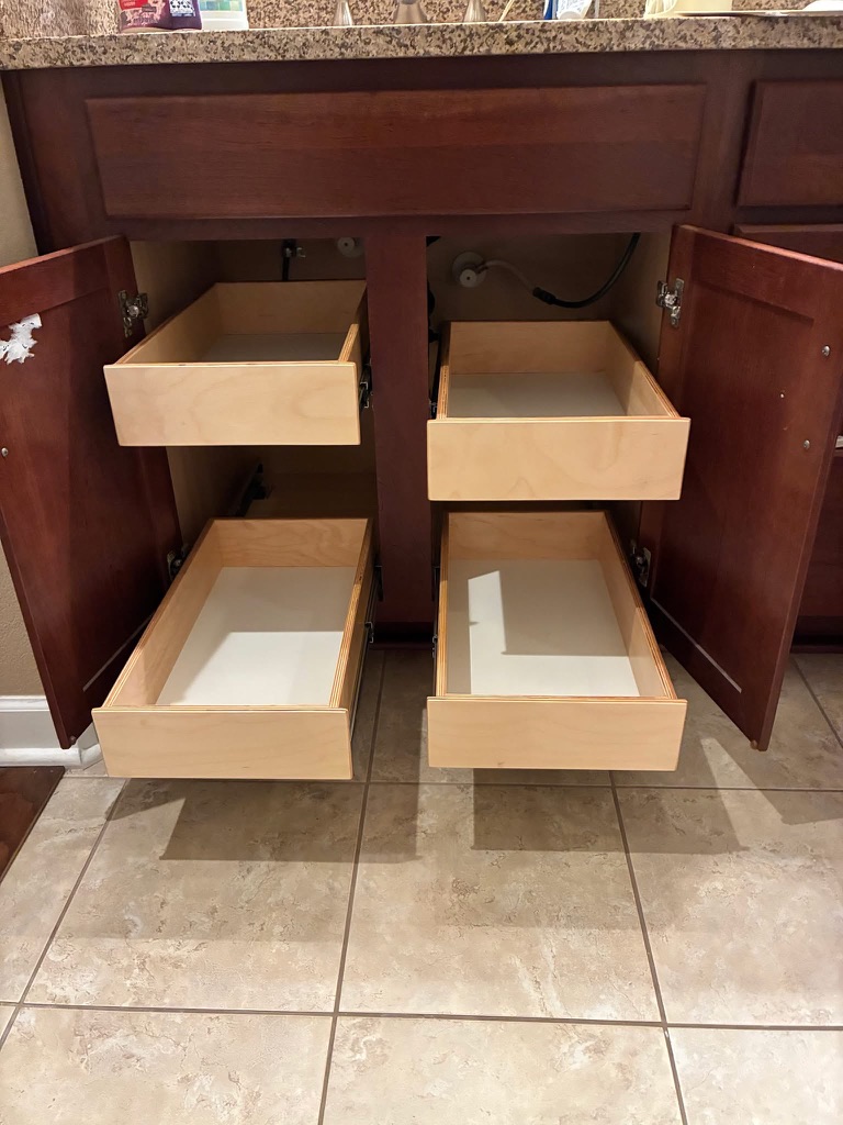 bathroom organization Four wooden drawers are pulled out from a bathroom vanity beneath a sink, with the cabinet doors open and plumbing visible. The drawers and cabinet are empty, and the floor is tiled.