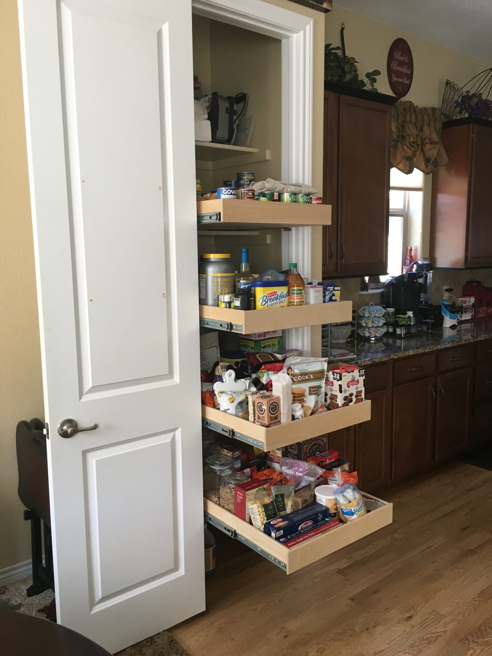 closet pantry approved for site A white pantry door is open, revealing four pull-out shelves filled with various food items and groceries in a kitchen with wooden cabinets and a hardwood floor.