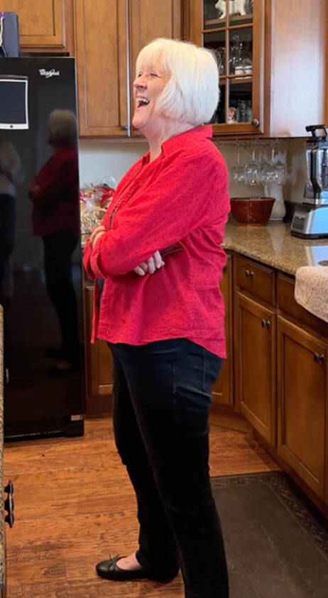 consultation-gallery-13 An older woman with short white hair, wearing a red shirt and black pants, stands smiling with arms crossed in a warmly lit kitchen with wooden cabinets and granite countertops.