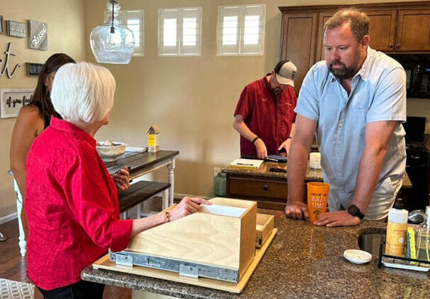 consultation-gallery-2 Four people are in a kitchen. An older woman in red talks to a man in blue leaning on the counter. Two others prepare food in the background. Kitchen counters have various items and wood boards. The atmosphere is casual and relaxed.