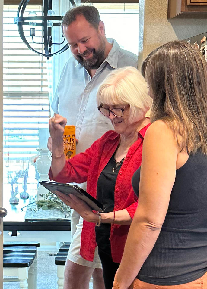 consultation-gallery-3 An older woman wearing glasses and a red shirt smiles and raises her fist while holding a tablet, standing with two other adults in a bright, sunlit kitchen.