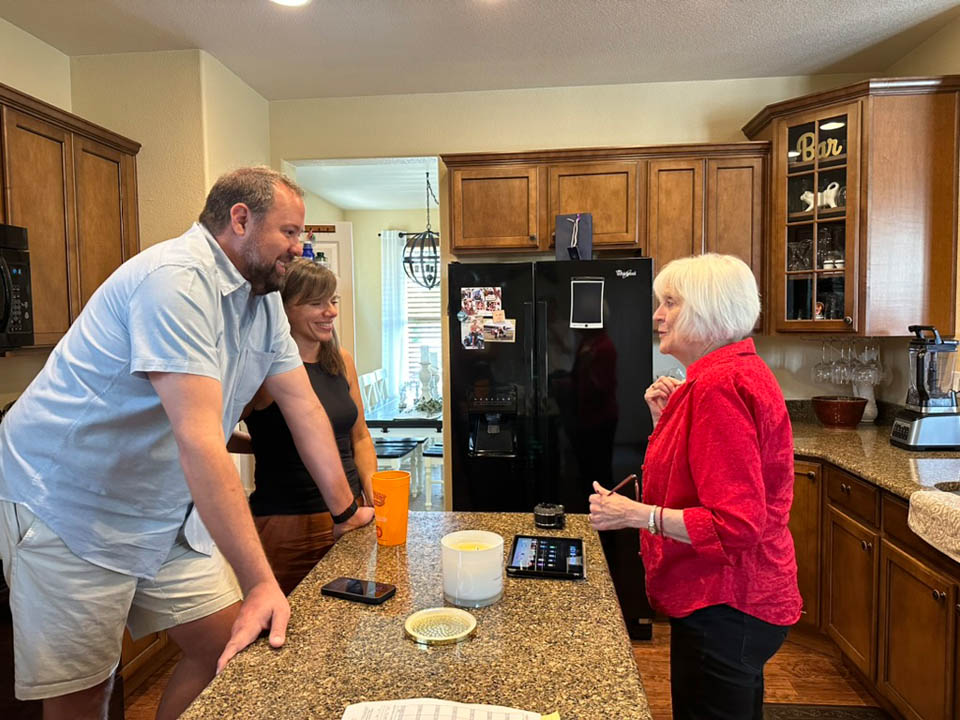 consultation-gallery-7 Three people are gathered in a kitchen, talking around a granite island countertop. A man and woman stand on one side, while an older woman in red stands on the other. Kitchen cabinets and appliances are visible in the background.