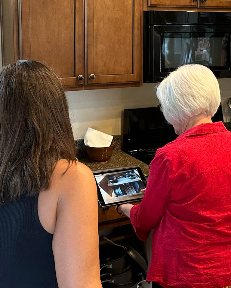 consultation-gallery-8 Two women stand in a kitchen, one with white hair and a red shirt using a tablet, while the other with brown hair in a black top looks on. Wooden cabinets, a microwave, and kitchen items are in the background.