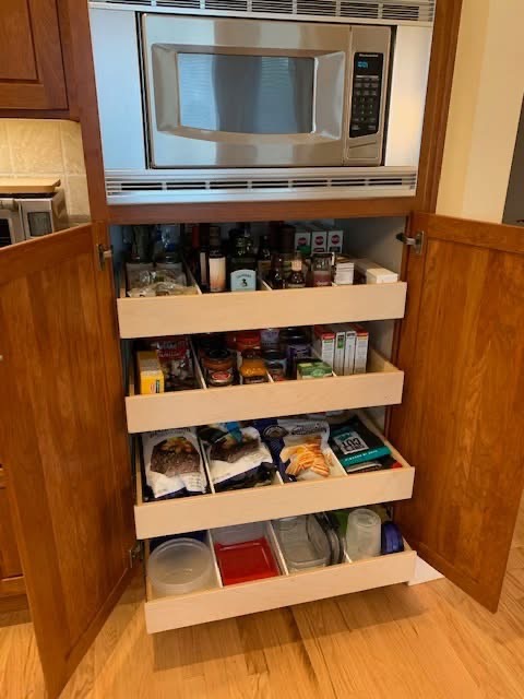 custom built under micro A kitchen cabinet with open wooden doors reveals several pull-out shelves stocked with pantry items, snacks, containers, and a microwave oven installed above the shelves.