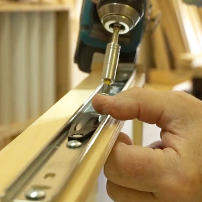 A close-up of a person’s hand installing a metal drawer slide onto a wooden board using a power drill in a woodworking shop.