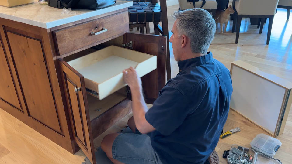 installation-gallery-1 A man is kneeling on the floor, installing or repairing a pull-out shelf inside a wooden kitchen cabinet. Tools and hardware are scattered nearby, and chairs and a dog are visible in the background.