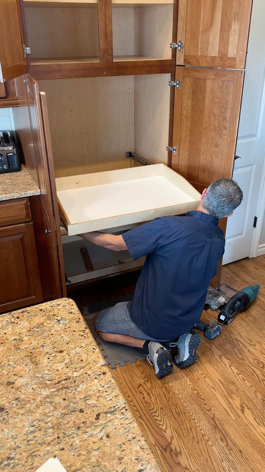 installation-gallery-10 A man kneels on a wooden floor, installing a sliding shelf inside a large wooden kitchen cabinet. Tools, including a power drill, lie nearby on the floor. The kitchen has brown cabinets and a granite countertop.