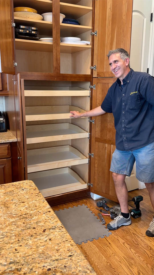 installation-gallery-17 A man in casual clothes stands in a kitchen next to a tall wooden cabinet with open doors and newly installed pull-out shelves. Tools and a gray mat are on the floor nearby.