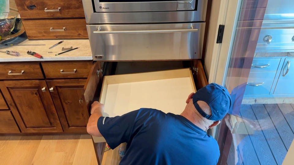 installation-gallery-18 A person in a blue shirt and cap installs or repairs a large pull-out drawer beneath a stainless steel oven in a wooden kitchen. Tools are visible on the nearby counter.