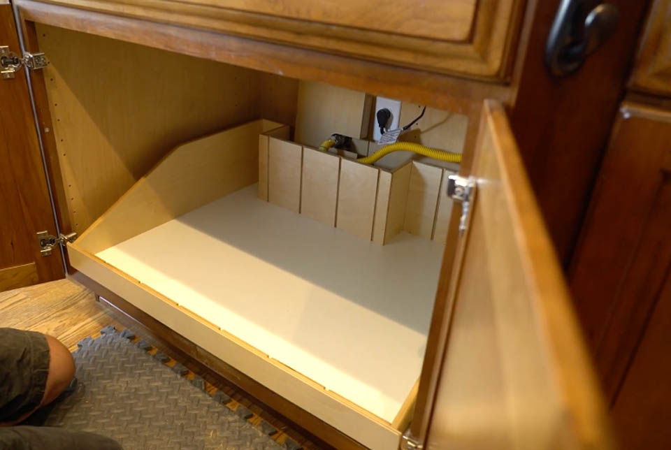installation-gallery-19 An open wooden cabinet under a kitchen counter reveals a custom-built wooden organizer with vertical dividers and a white base. A yellow gas line, valve, and an outlet are visible at the back.