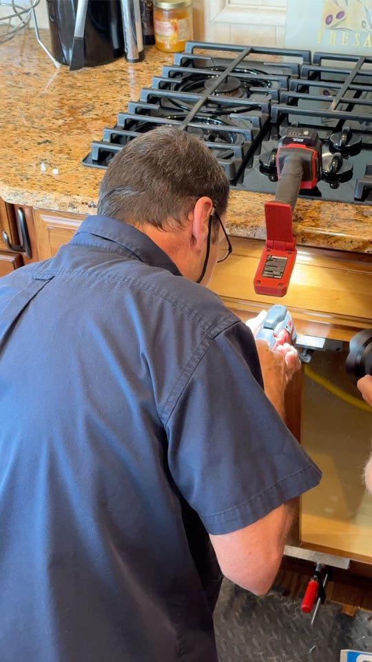 installation-gallery-4 A man in a navy shirt works under a kitchen stove, using a tool to repair or install something inside a cabinet. Cooking grates and a clamp are on the countertop above him.