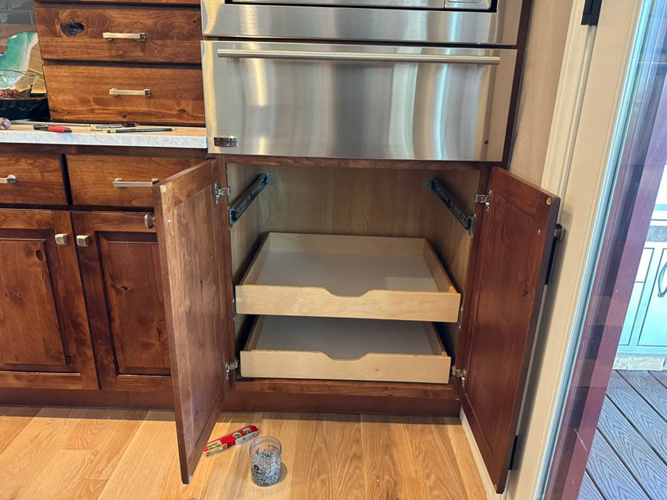 installation-gallery-6 An open kitchen cabinet with two pull-out wooden shelves. The cabinet doors are open, and tools are on the floor nearby, indicating installation or repair work. The kitchen has wood cabinets and a stainless steel appliance above.