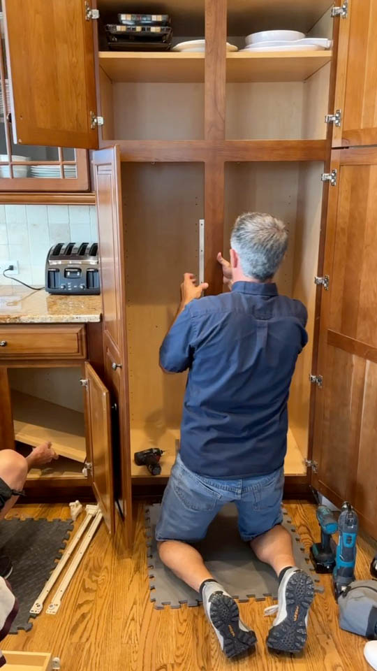 installation-gallery-7 A man kneels on a foam mat, measuring the inside of a large wooden kitchen cabinet with a tape measure. Tools and cabinet parts are scattered nearby on the hardwood floor.