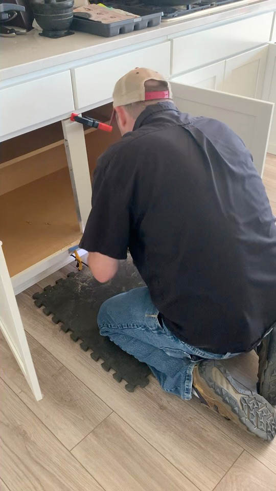 installation-gallery-9 A person wearing a cap, jeans, and work boots kneels on a foam mat while working on the inside of a kitchen cabinet with the doors open. Tools and supplies are visible nearby on the counter.