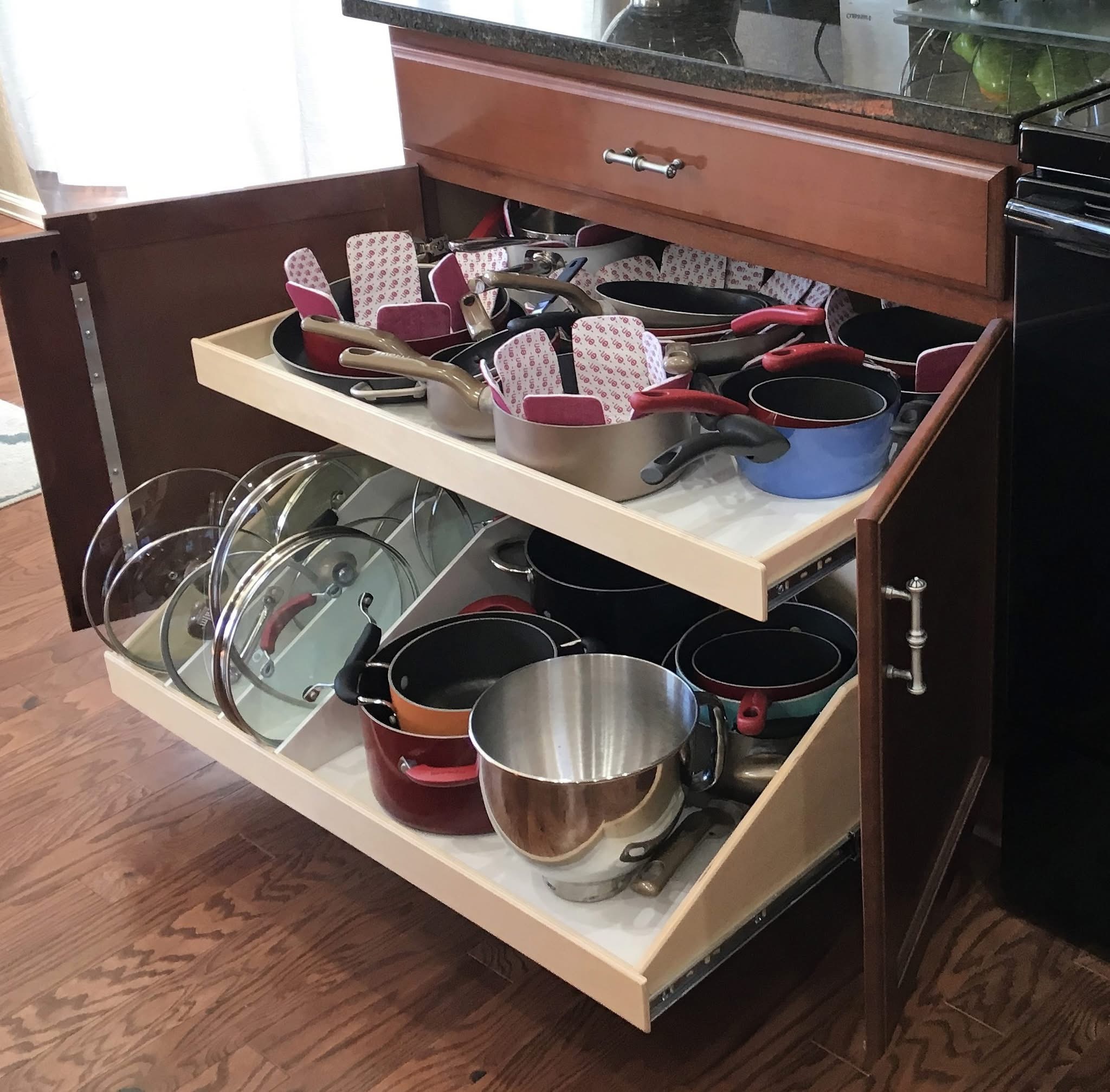 maximum pots and pan storage Open kitchen cabinet with two pull-out shelves neatly organized with pots, pans, lids, and baking trays. The shelves are wooden, and the cabinet is brown with metal handles. The kitchen floor is hardwood.