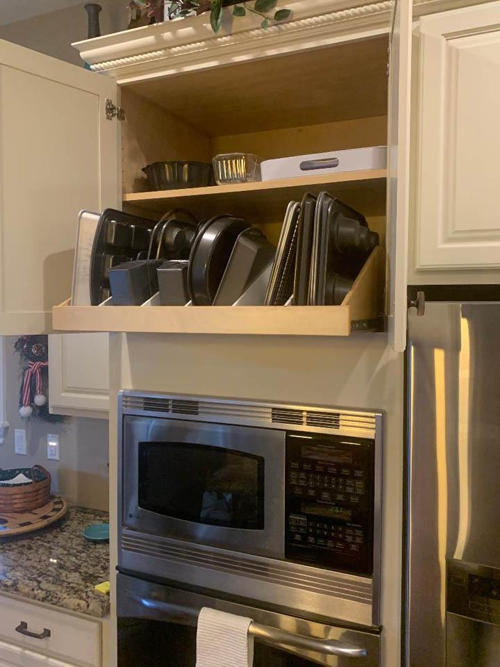 reos-product-gallery-1 A kitchen cabinet above a built-in microwave oven stores various baking pans and trays organized vertically on a pull-out shelf. The cabinet doors are open, showing neatly arranged bakeware.