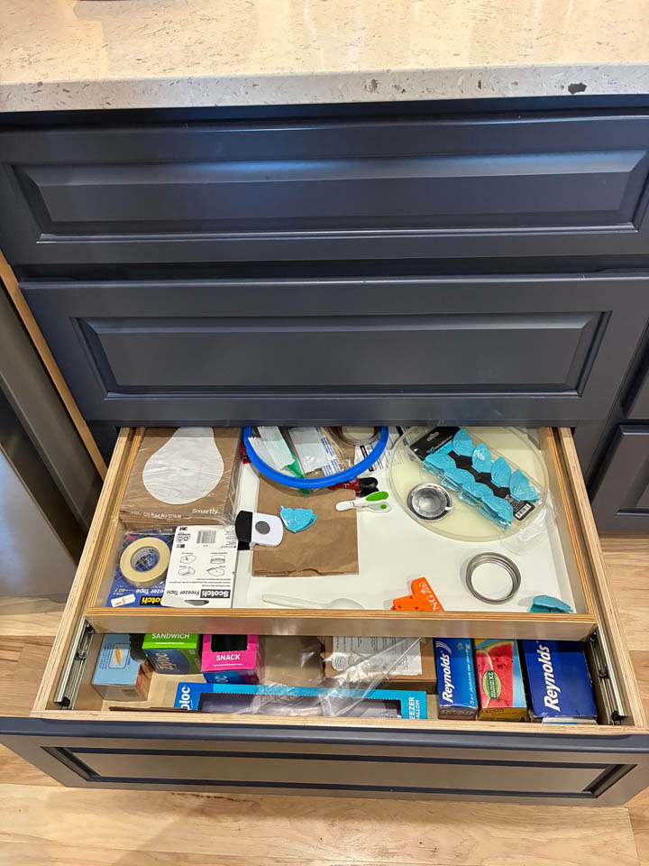 reos-product-gallery-13 A kitchen drawer partly open, containing various items such as tape, scissors, plastic wrap, ziplock bags, pens, and other miscellaneous supplies, with dark cabinets and a light countertop above.