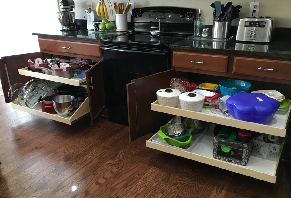 reos-product-gallery-15 Kitchen with wooden cabinets and sliding drawers open, showing organized storage of pots, pans, baking sheets, and plastic food containers. Black appliances, utensils, and small kitchen gadgets are on the counter.