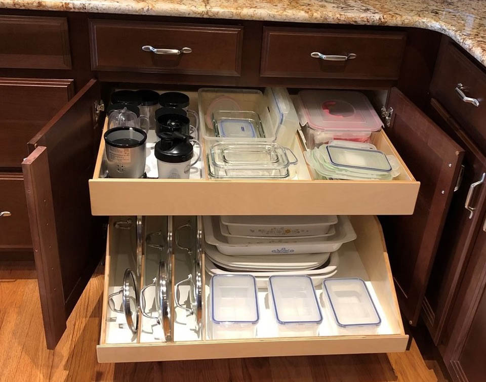 reos-product-gallery-18 Two pull-out kitchen drawers organized with glass containers, plastic lids, baking dishes, blender cups, and metal pot lids. The drawers are open, showing efficient storage in a wooden cabinet.