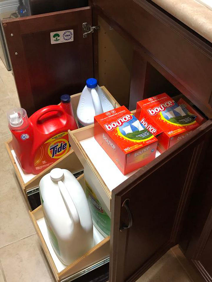 reos-product-gallery-19 A wooden cabinet with pull-out shelves holding laundry supplies, including Tide detergent, Bounce dryer sheets, bleach, and other cleaning products, neatly organized.