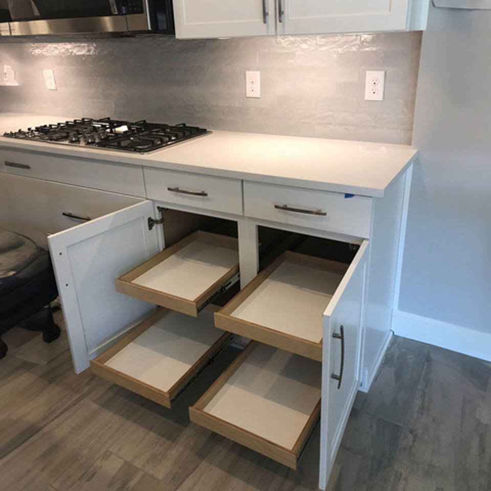 reos-product-gallery-2 A modern kitchen with a gas stove, white cabinets, and two lower cabinet doors open to reveal pull-out wooden shelves on both sides. The floor is light-colored wood, and the walls have a light gray backsplash.
