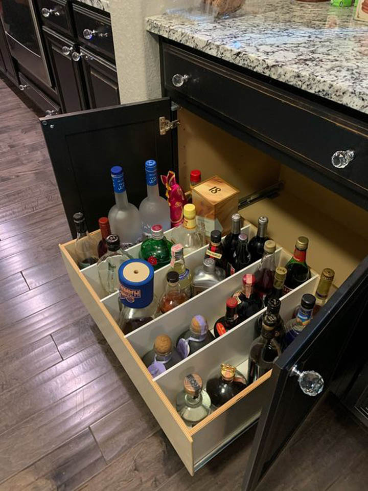reos-product-gallery-20 A kitchen cabinet drawer is pulled out to reveal organized bottles of various alcohol, including vodka, whiskey, and wine, neatly stored upright in divided compartments below a granite countertop.