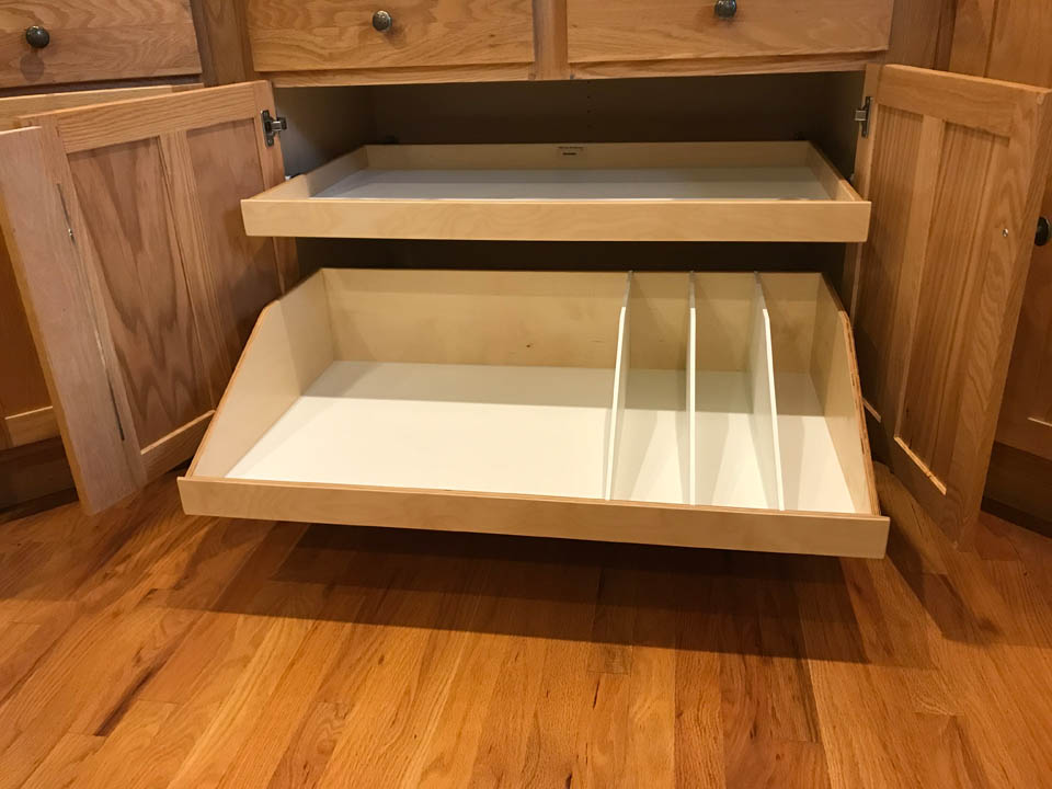 reos-product-gallery-21 A wooden kitchen cabinet with two pull-out shelves; the bottom shelf has vertical dividers creating separate compartments. The cabinet doors are open, revealing the organized storage inside.