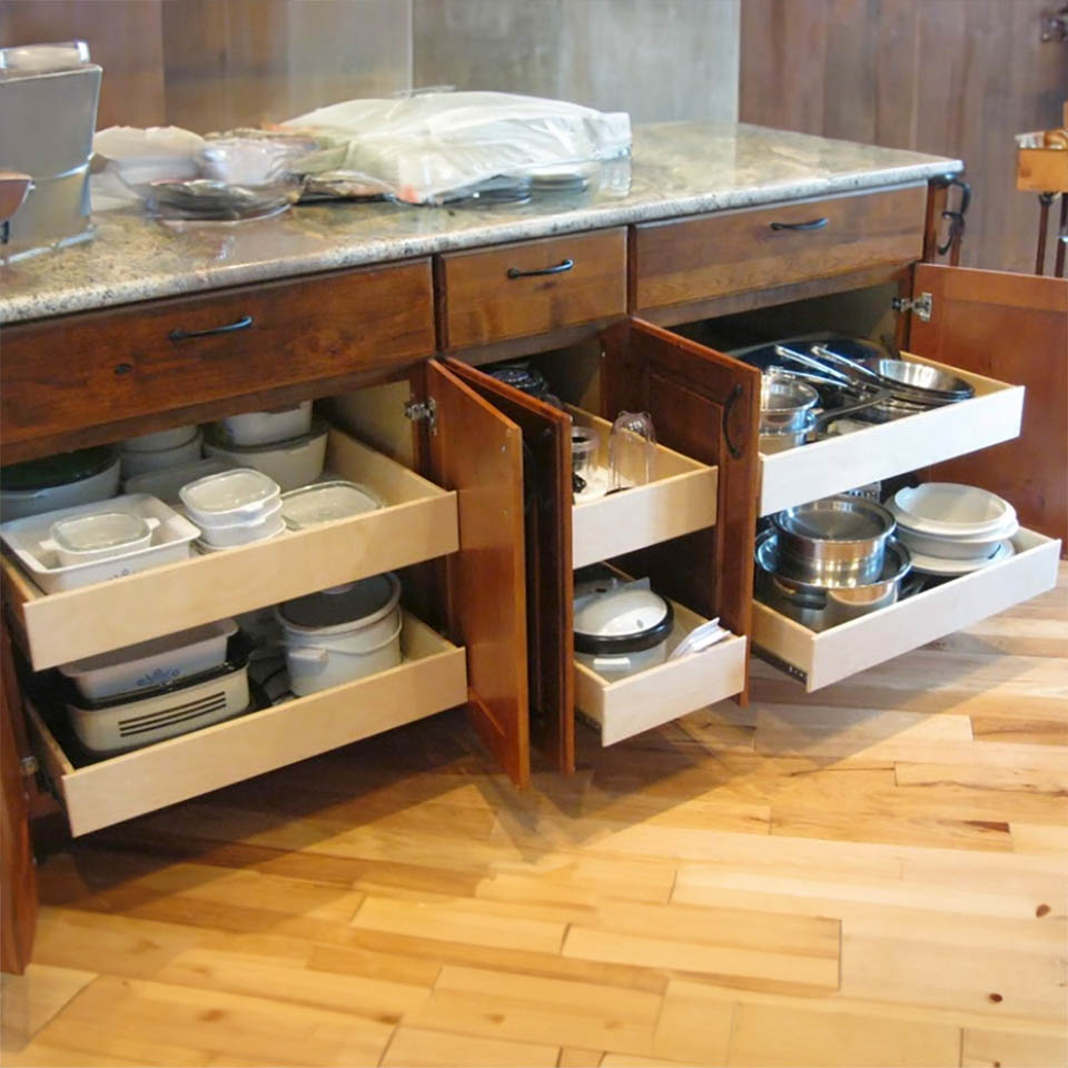 reos-product-gallery-27 Open wooden kitchen cabinets with several pull-out drawers, neatly organized with containers, lids, pots, and pans under a granite countertop on a wooden floor.