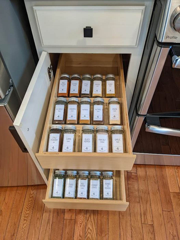 reos-product-gallery-28 A kitchen drawer slides open to reveal two tiers filled with neatly organized glass spice jars, each labeled with the spice name. The drawer is next to an oven and has wooden shelves and a white front panel.