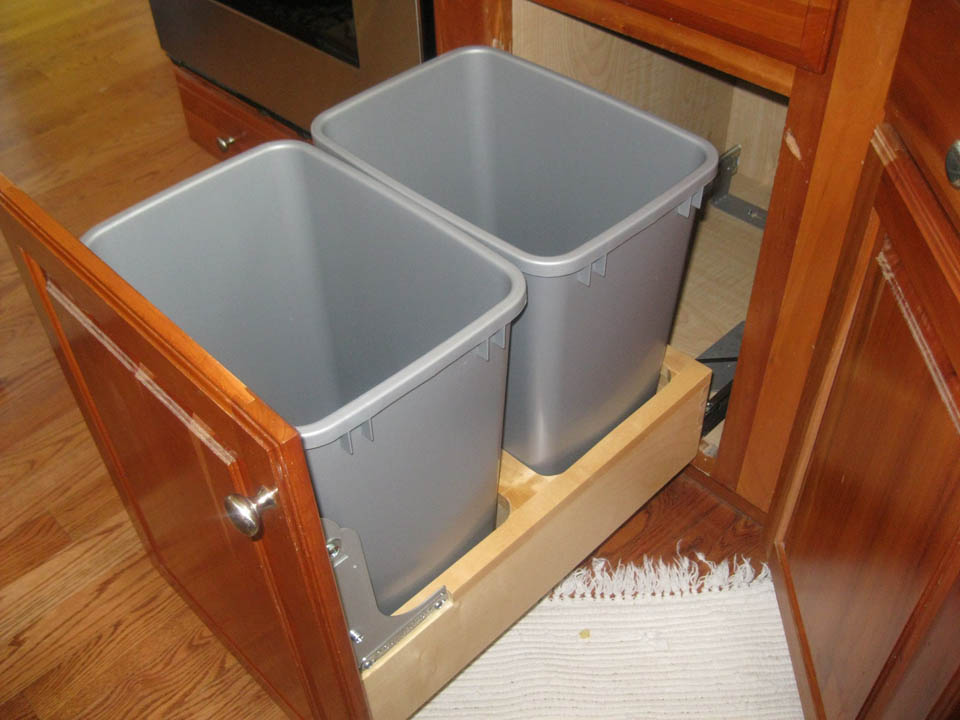 reos-product-gallery-29 Two gray plastic trash bins are placed side by side in a wooden pull-out drawer inside a kitchen cabinet, with part of a white rug visible on the wooden floor in front.