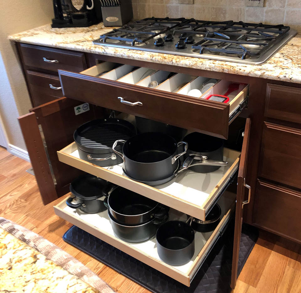 reos-product-gallery-30 A kitchen cabinet with sliding drawers open, showing neatly organized pots, pans, and lids below a countertop with a gas stove and coffee maker.