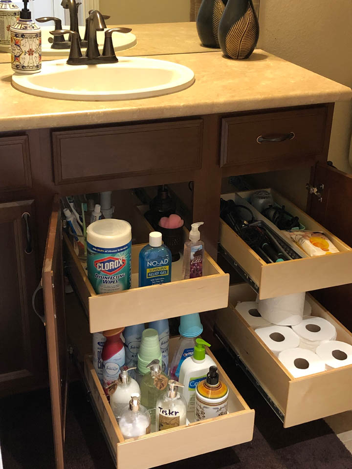 reos-product-gallery-6 Bathroom vanity with open drawers and cabinets showing organized cleaning supplies, toiletries, hand soap, and toilet paper rolls stored neatly on pull-out shelves below a sink.