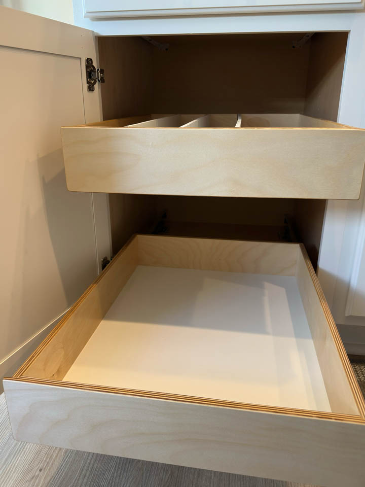 reos-product-gallery-7 Open kitchen cabinet showing two wooden pull-out drawers; the top drawer has divider sections, while the lower drawer is open and empty. The cabinet interior is light wood, and the floor is light-colored.