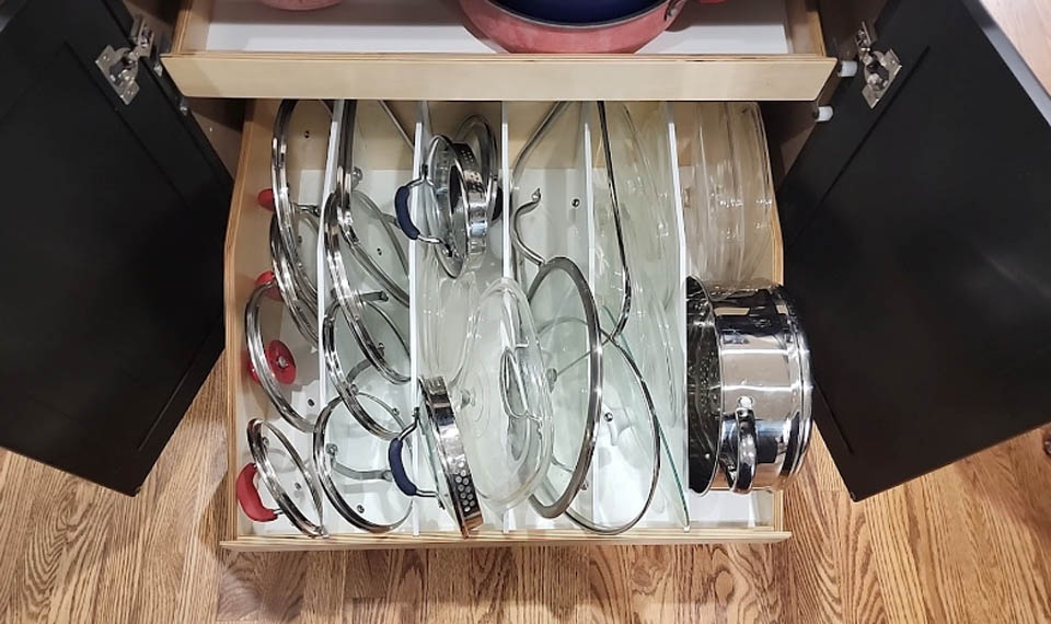 reos-product-gallery-9 A kitchen drawer with vertical dividers neatly organizing various pot and pan lids, with some metal pots and pans on the right. The drawer is open, revealing a wood floor and black cabinet doors.