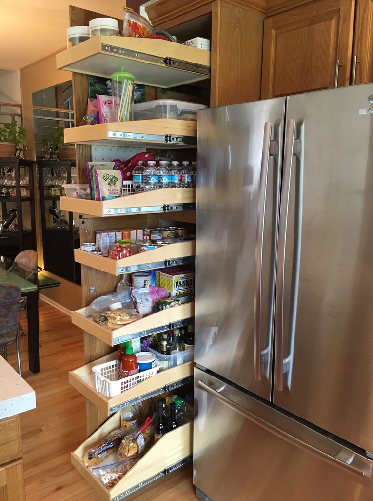 retro for deep tight spaces A tall wooden pull-out pantry with six shelves is fully extended next to a stainless steel refrigerator, showing various organized food items, condiments, bottled water, and snacks on each shelf.