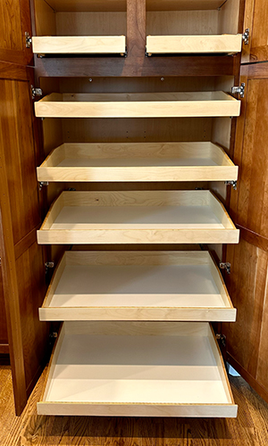 A wooden pantry cabinet with its doors open, revealing six empty sliding shelves made of light-colored wood, all pulled out to different lengths. The cabinet sits on a hardwood floor.