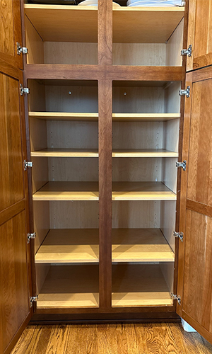 An empty wooden pantry cabinet with double doors is open, revealing six shelves inside. The wood has a warm brown finish, and the shelves and interior have a lighter color. The floor beneath is hardwood.